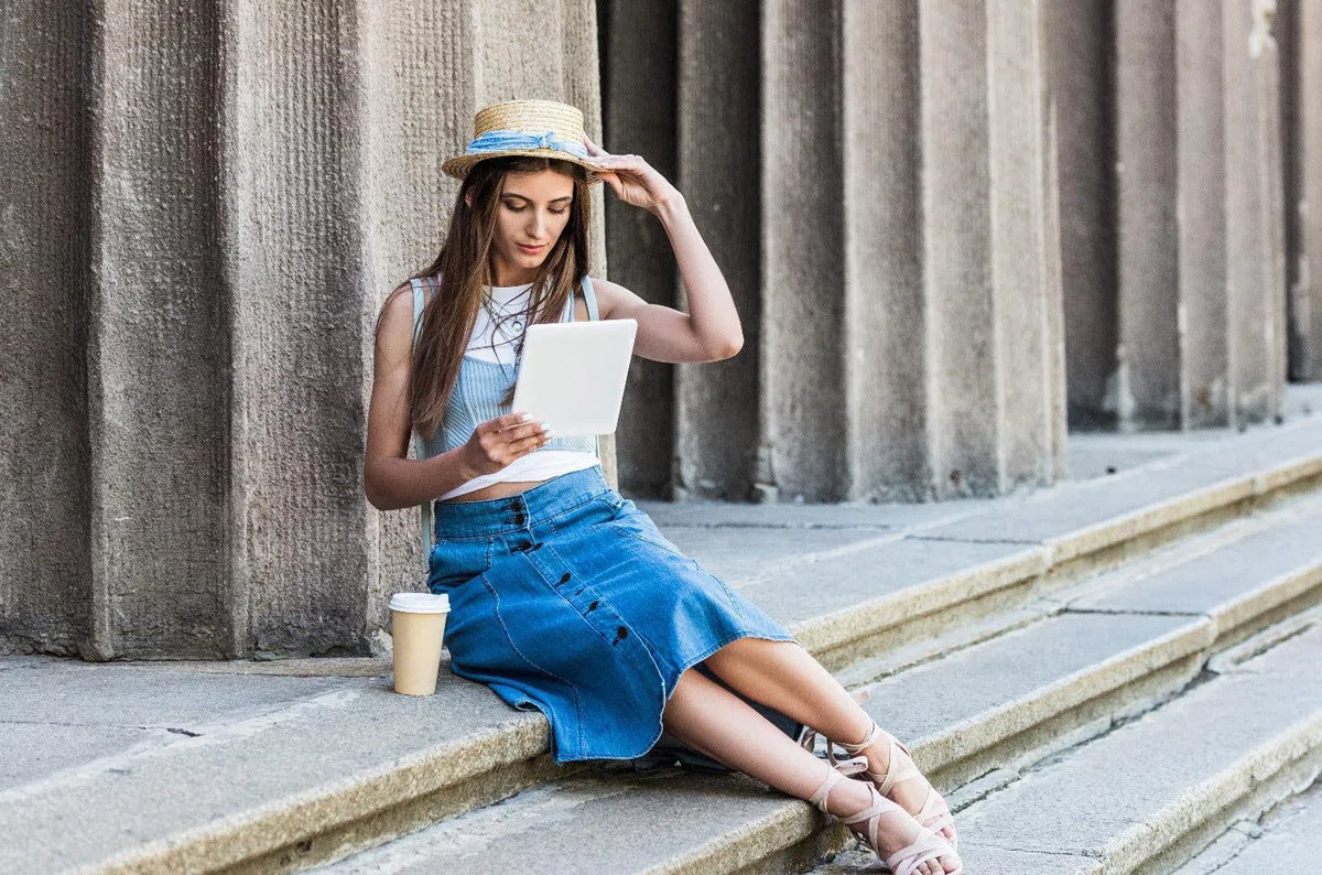 A women in long jean skirt sitting on stairs with a computer and a cup of coffee.